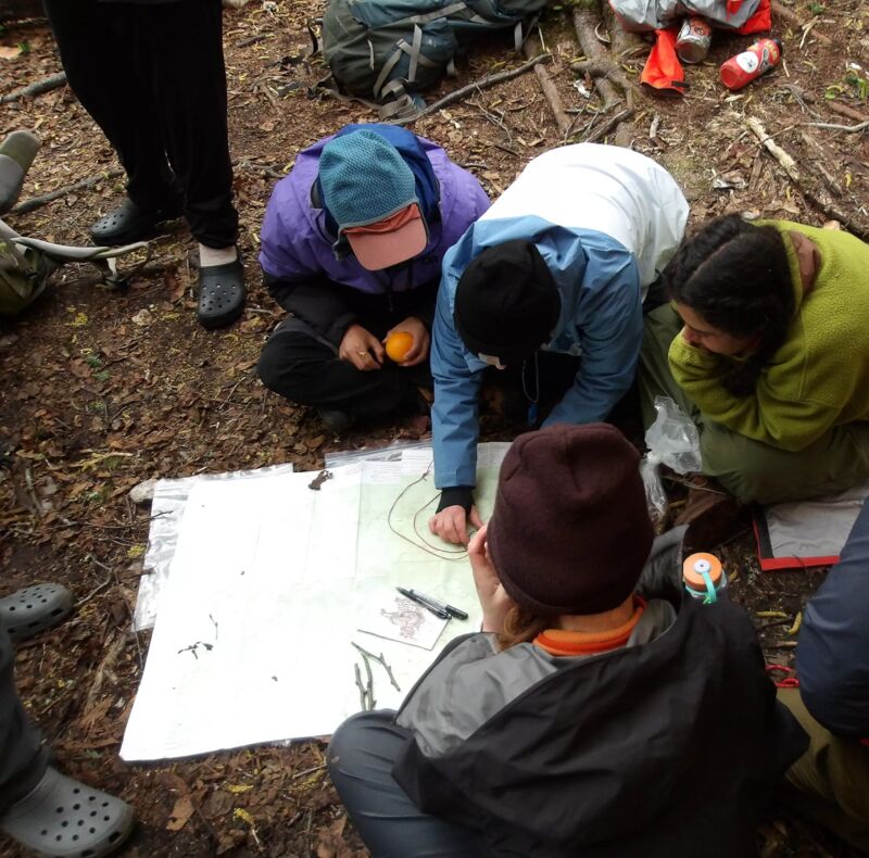A group of people are gathered around a map spread out on the ground in a wooded area. They appear to be planning or studying the map, possibly for navigation or a similar purpose. Some individuals are pointing at the map, and there are backpacks and other items scattered around, suggesting they may be hiking or camping.